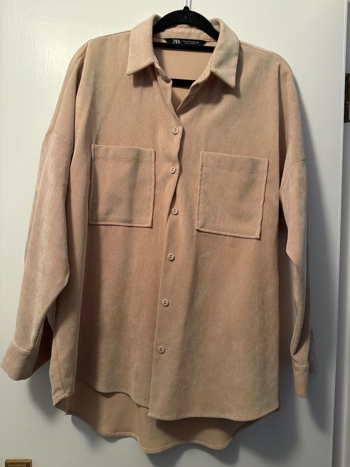 Beige long-sleeved shirt with buttons and two front pockets, hanging on a black hanger over a white door.
