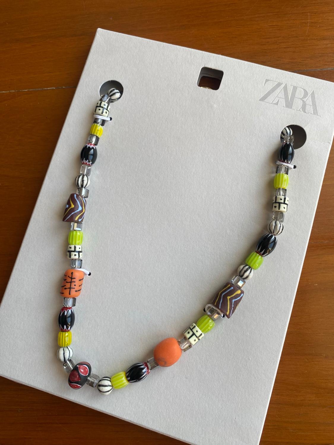 Coloured and differently shaped beaded necklace on white card with the text ZARA, on a wooden table.