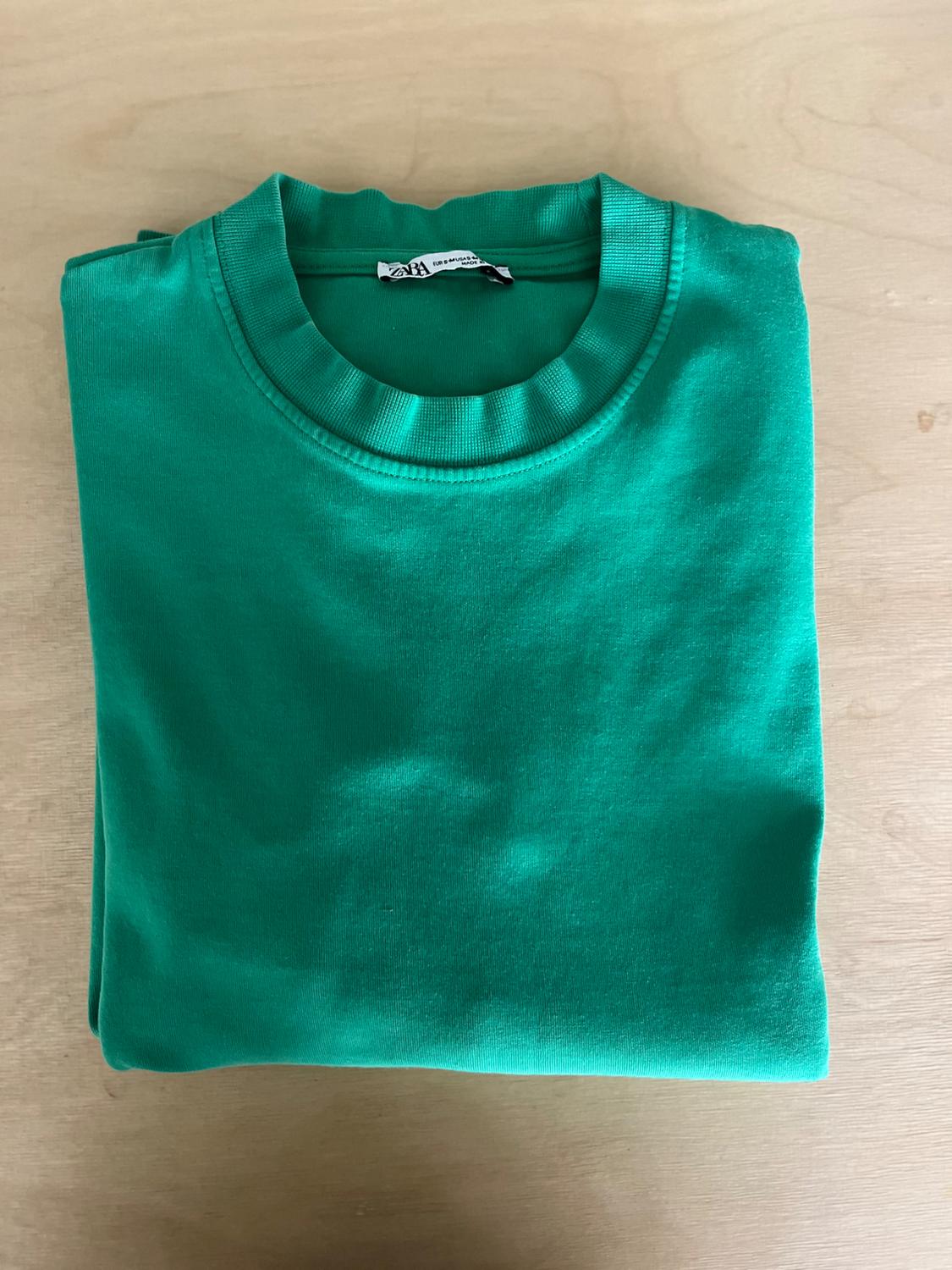 Green sweatshirt folded on a light wooden surface. The label is visible on the collar.