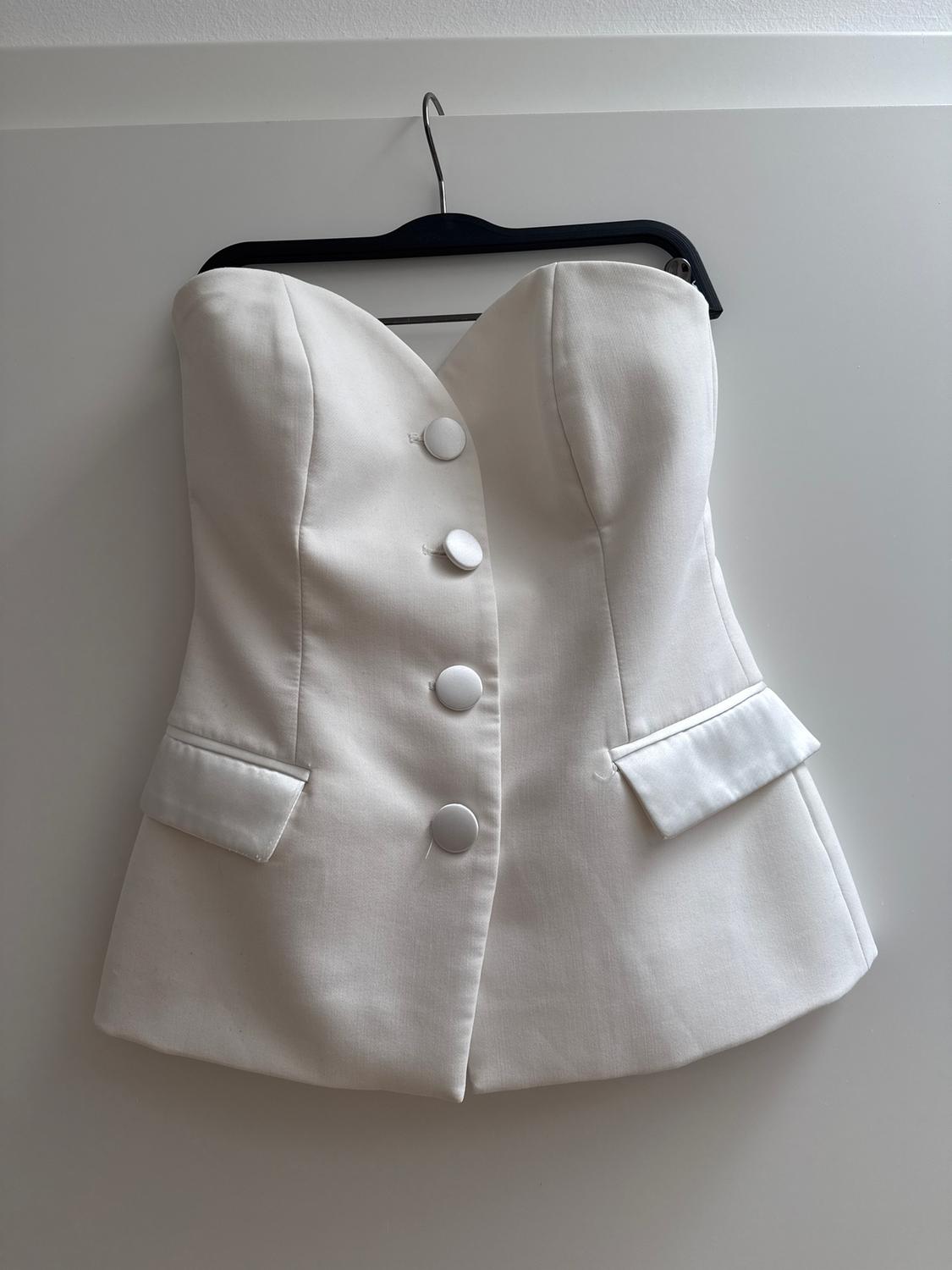 White strapless waistcoat with large buttons and pockets, hanging on a black hanger against a light grey background.