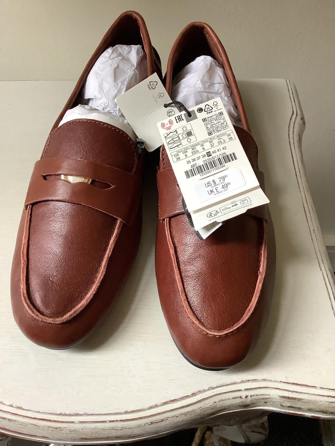 Brown leather loafers on a distressed white table. Label with text: ART: 2535/611/105 US $79.90 UK £49.99.