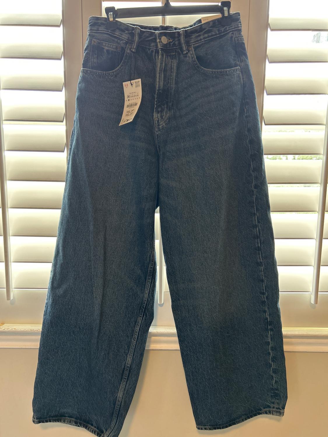 Blue denim jeans hanging in front of a window with white blinds. Label visible on the left front pocket.