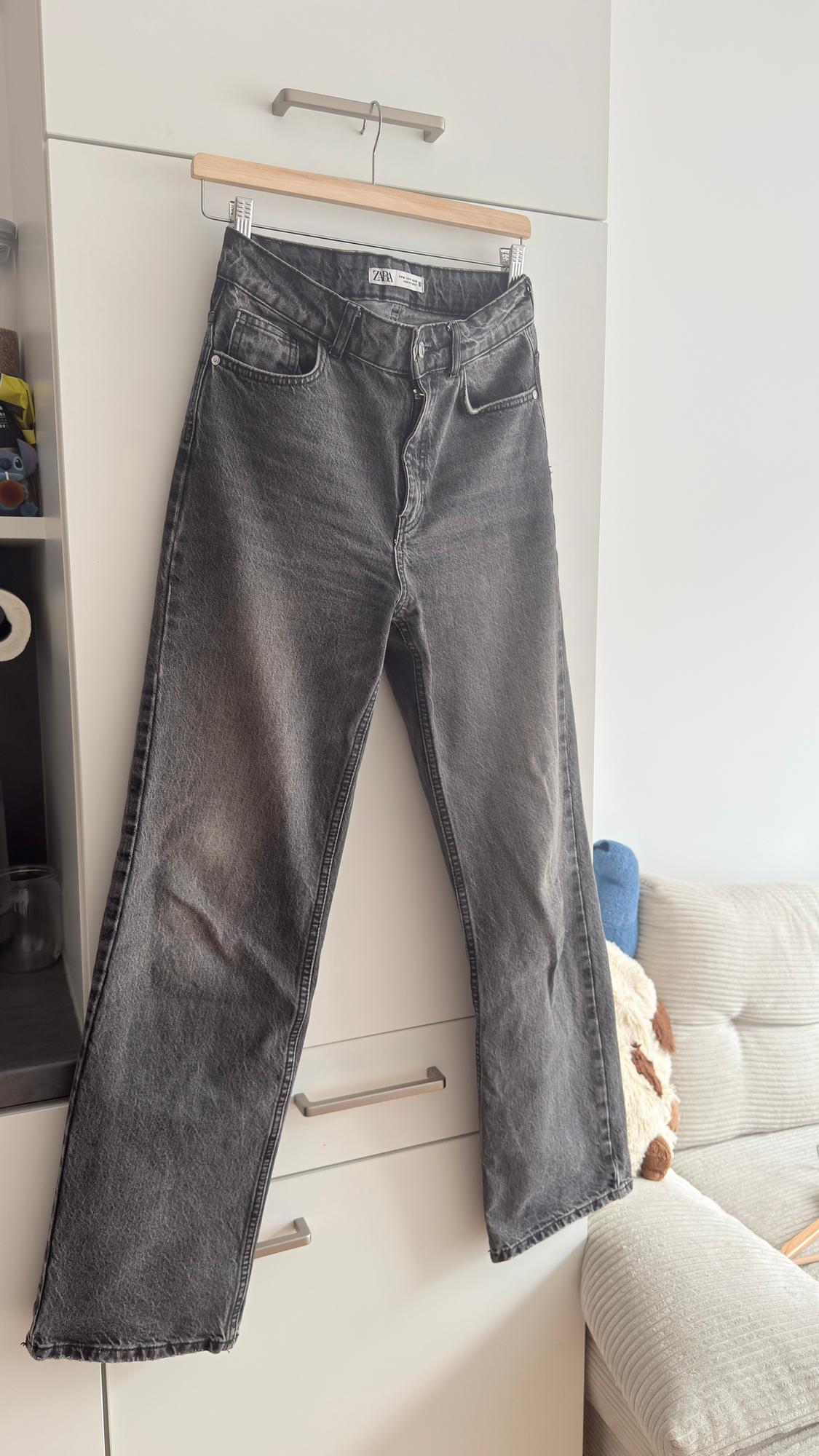 Grey jeans hanging on a wooden hanger above a white wardrobe, with a visible label on the waistband.