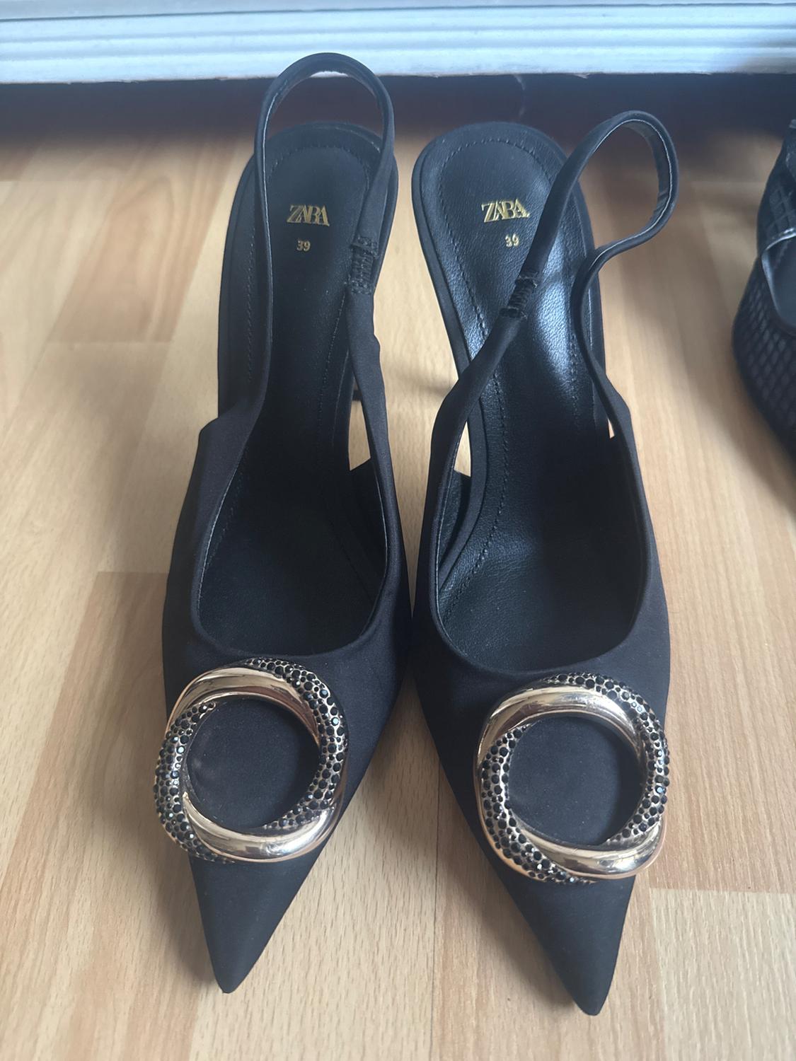 Black high-heeled shoes with a pointed toe and a gold ring embellishment on the front. Visible text: "ZARA 39".