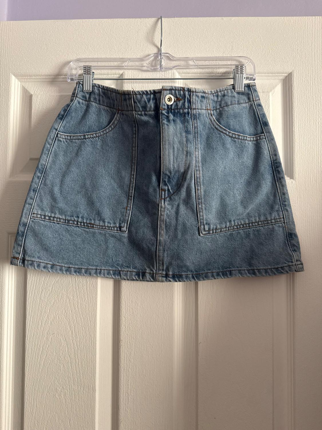 Light blue denim skirt hanging on a clear hanger over a white door. Design with front pockets and button.