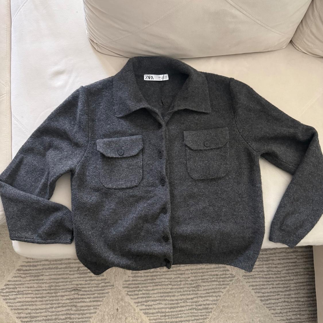 Dark gray knit jacket with collar, buttons, and pockets, on a white sofa and rug. Label with ZARA text.
