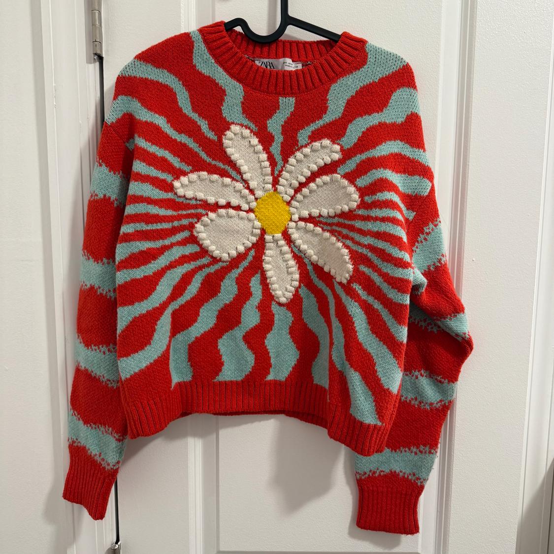 Red sweater with blue wavy stripes and a white daisy with a yellow center on the front, hanging on a white door.