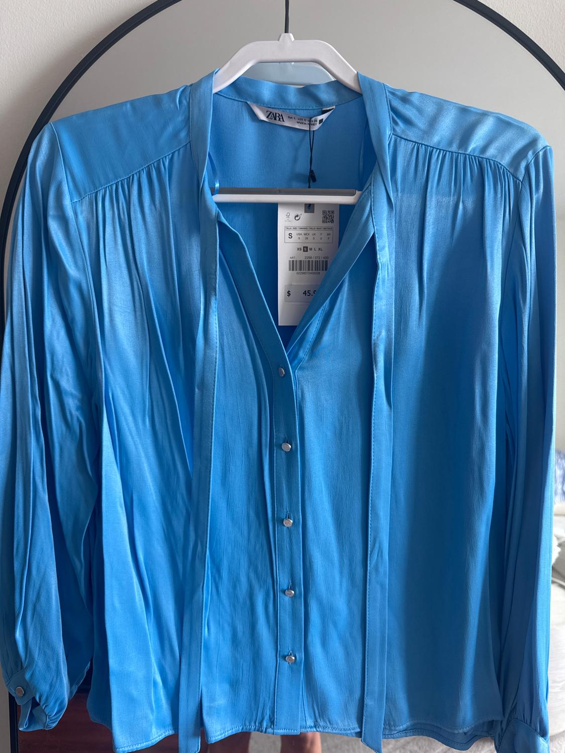 Blue long-sleeved blouse hanging on a white hanger in front of a mirror. Visible tag with price of £45.95.
