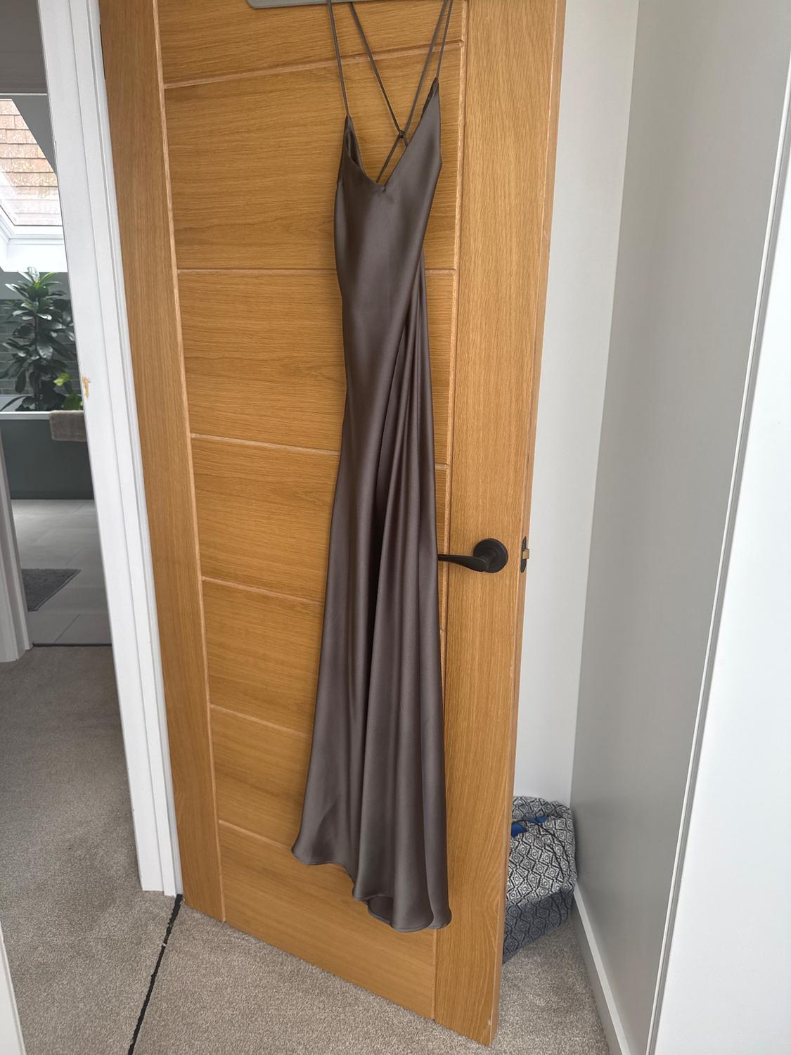 Long, thin-strapped grey dress hanging on a wooden door. Background with a beige rug and a potted plant.