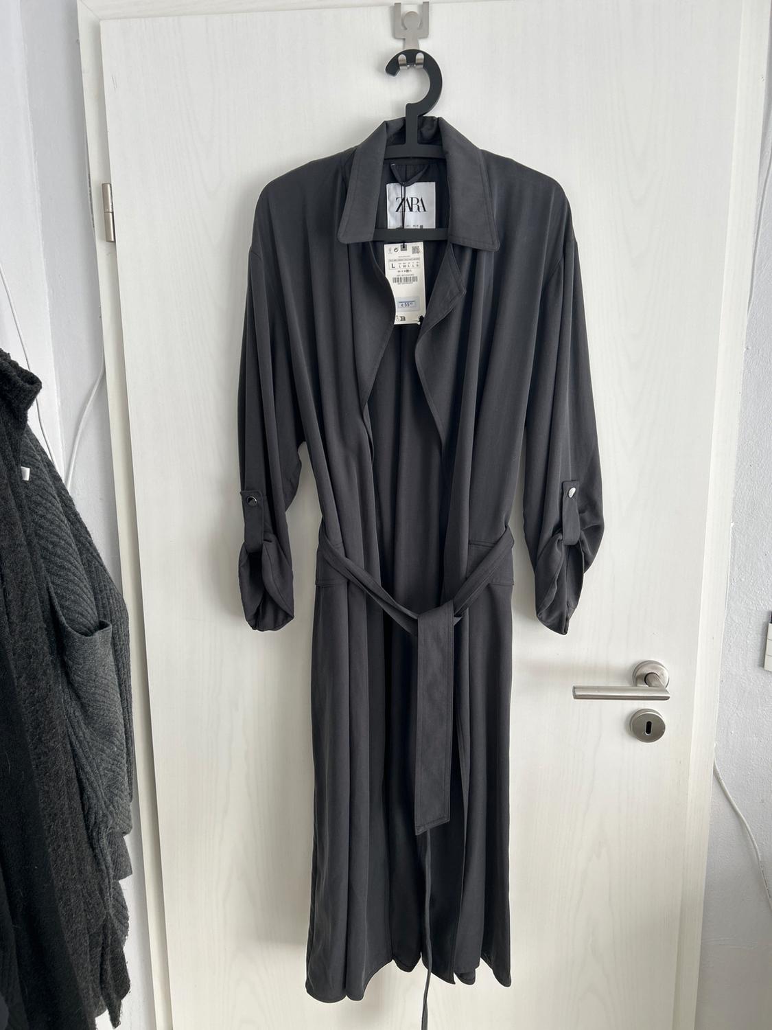 Black coat hanging on a white door, with a belt and a visible Zara label. Next to it, part of another dark garment can be seen.