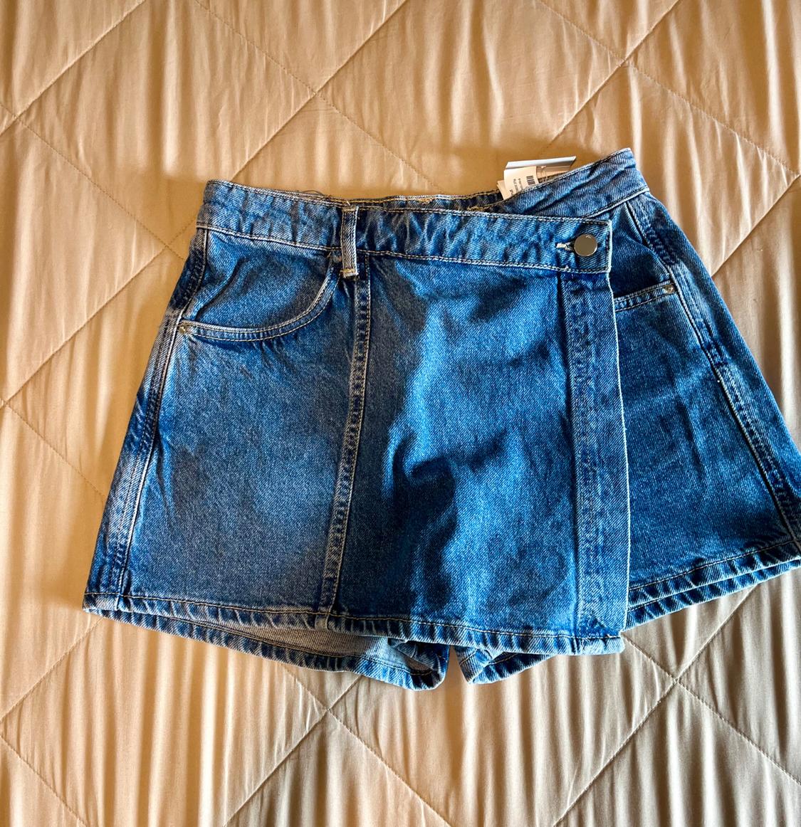 Blue denim skort with a crossover front panel and button, on a quilted beige comforter. Casual style.
