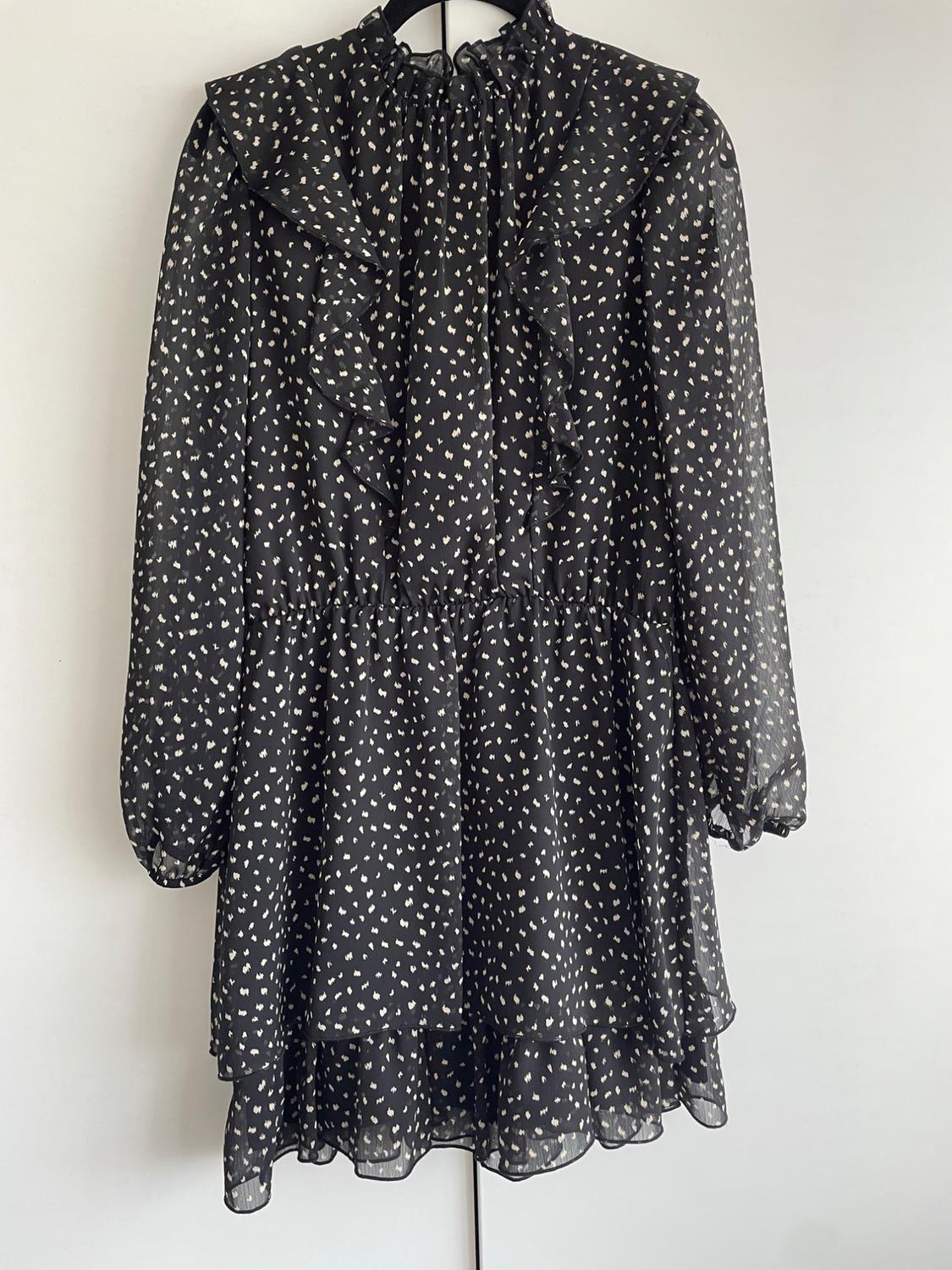 Black dress with a small white polka dot print, frills on the chest and long sleeves, hanging on a hanger against a white background.