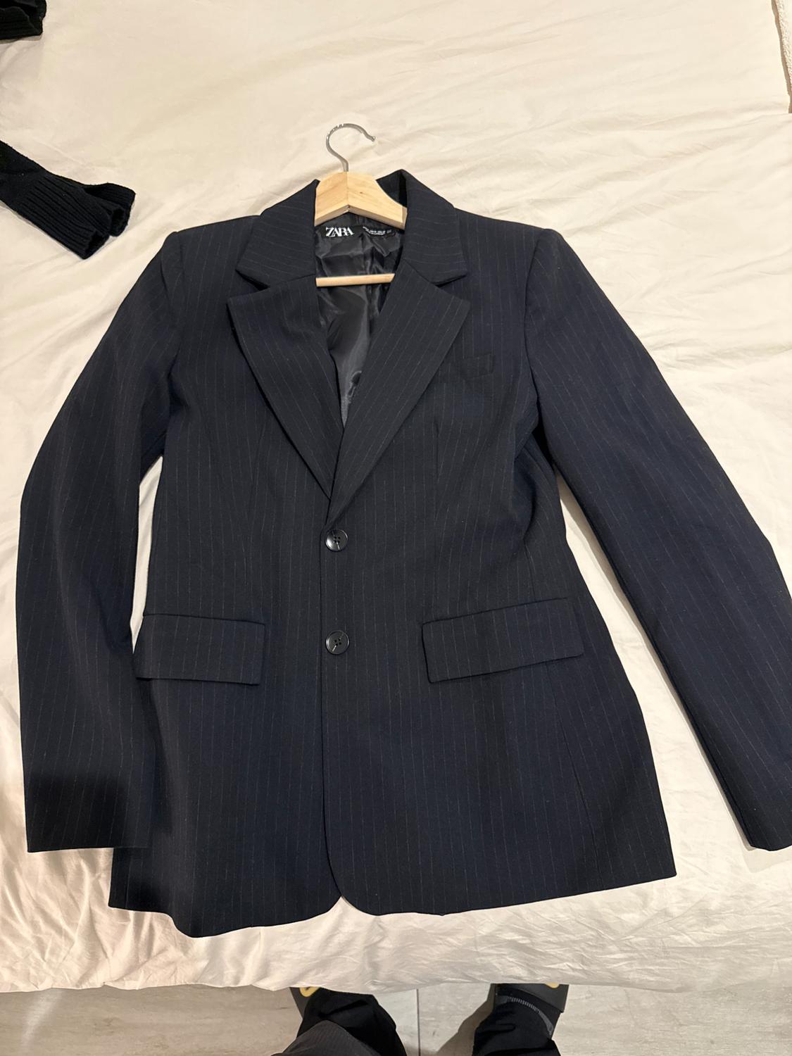 Black pinstripe jacket hanging on a wooden hanger over a white bed. Visible label with text "ZARA".