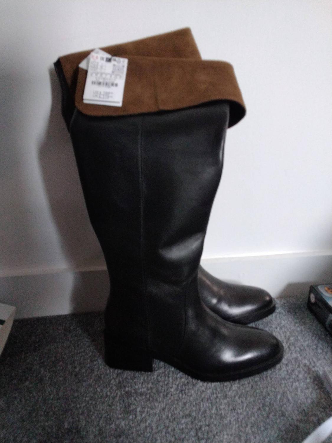 Black high boots with a folded-over brown top cuff and tag, on a grey carpet in front of a white wall.