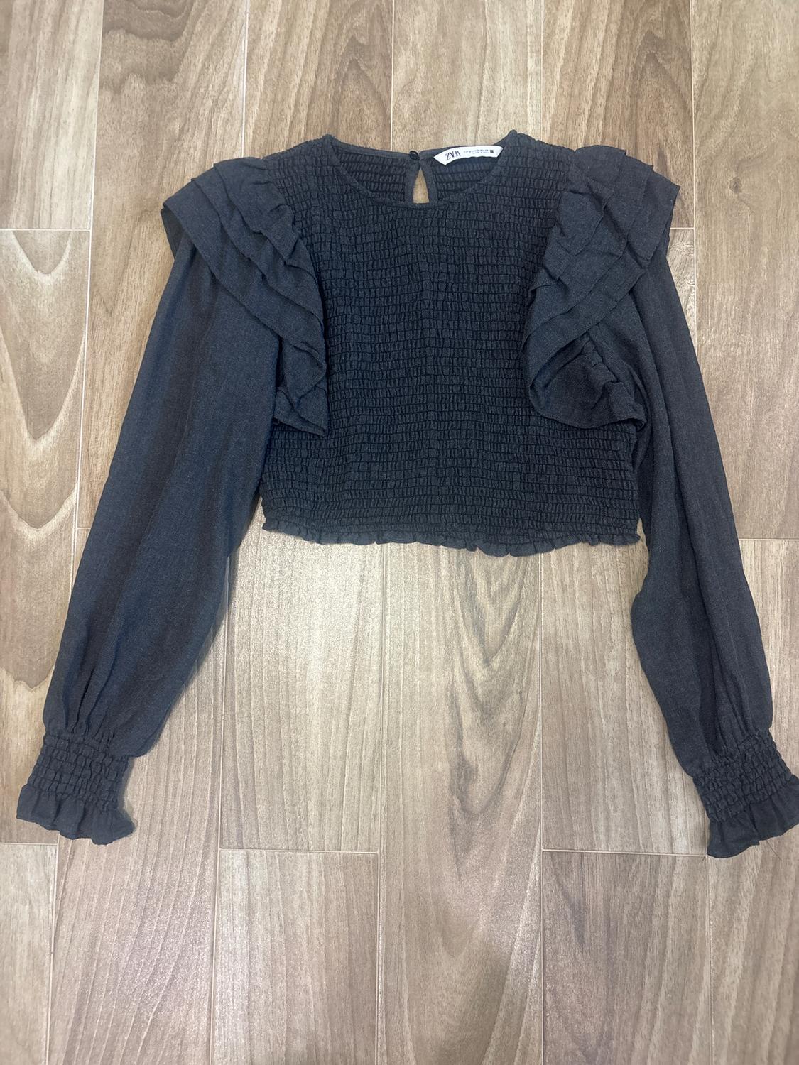 Black long-sleeved blouse with ruffled shoulders and ruched fabric, on a light wooden floor. Visible tag at the neck.