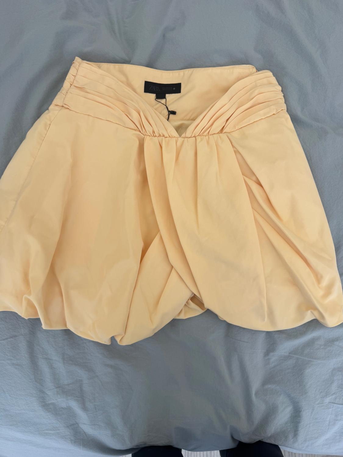Light yellow short skirt, ruched and puffed, on a blue-grey sheet. Black label at the waist with the text ZARA.