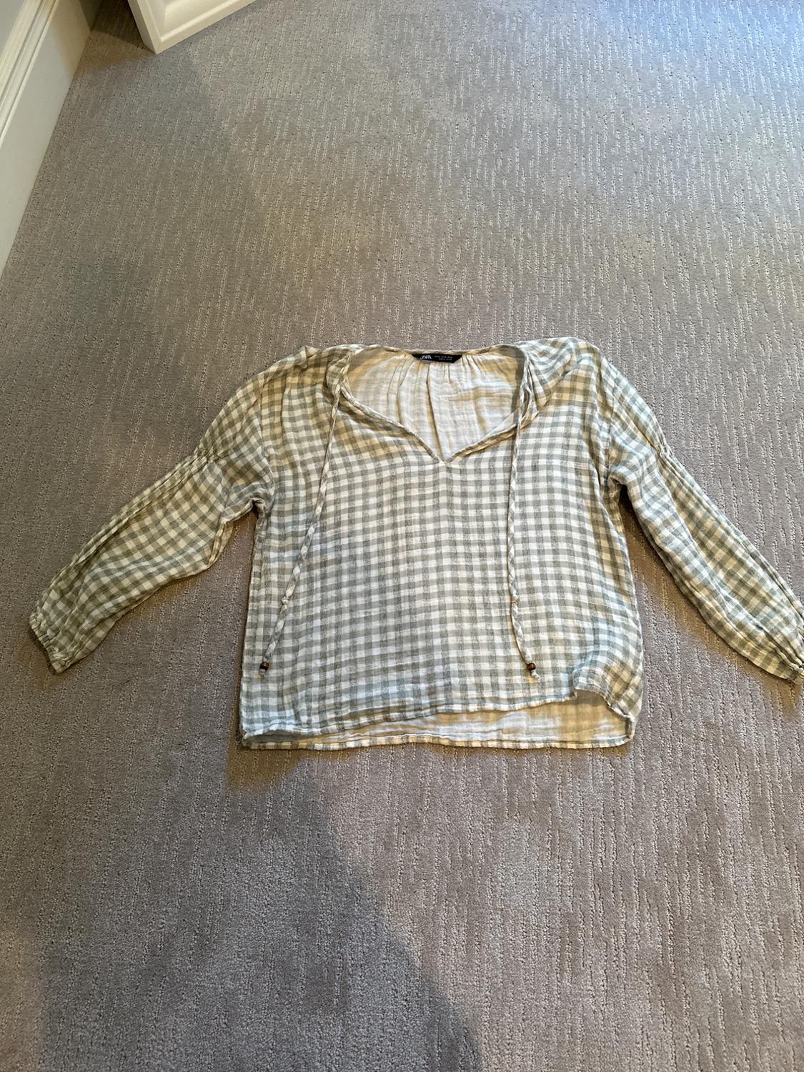Light-toned plaid shirt with a hood and drawstrings, laid out on a beige rug in an indoor setting.