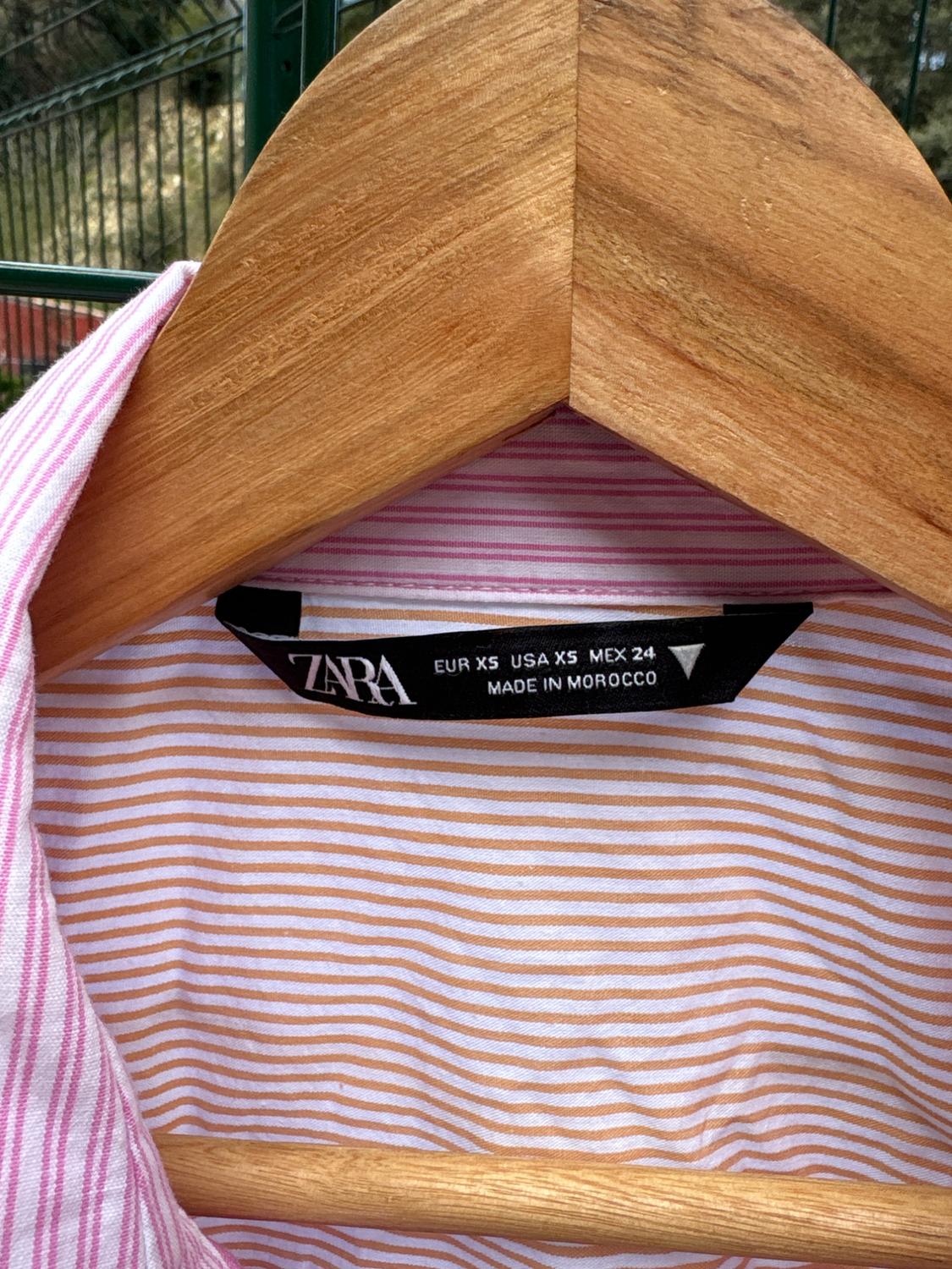 Pink and white striped shirt on wooden hanger. Black label: "ZARA, EUR XS, USA XS, MEX 24, MADE IN MOROCCO". Garden background.