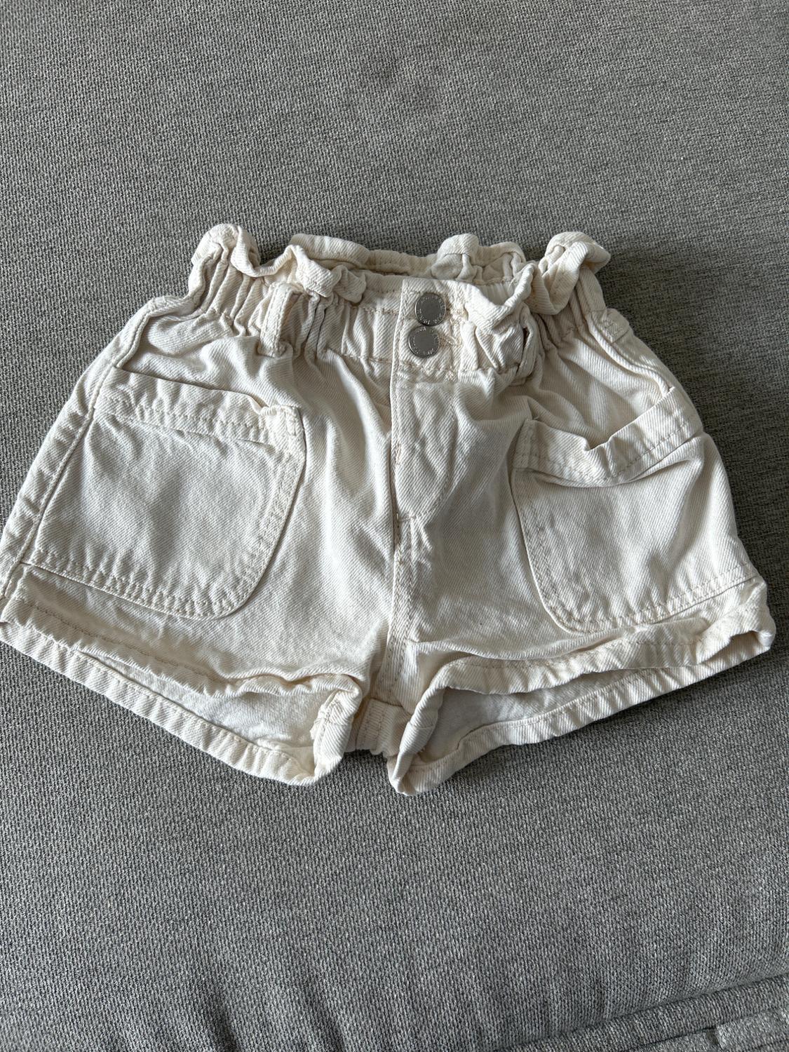 White denim shorts with a shirred elasticated waist and two buttons, on a textured grey background.