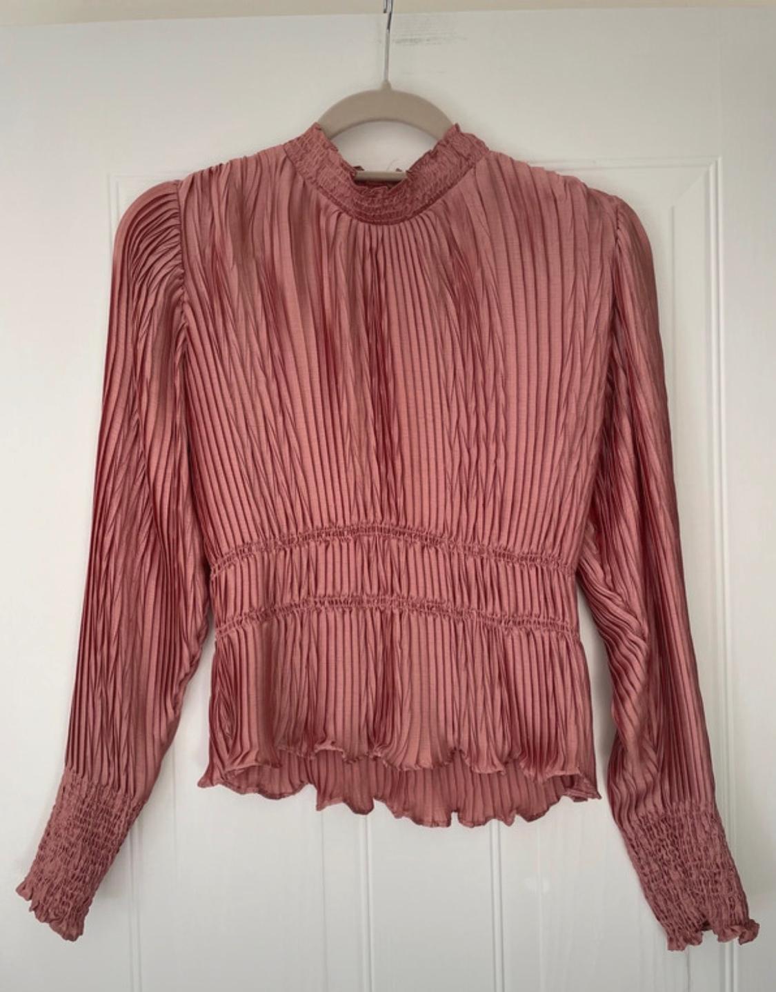 Pink pleated long-sleeved blouse with a high neck, ruffled cuffs and waistband, hanging on a hanger over a white door.