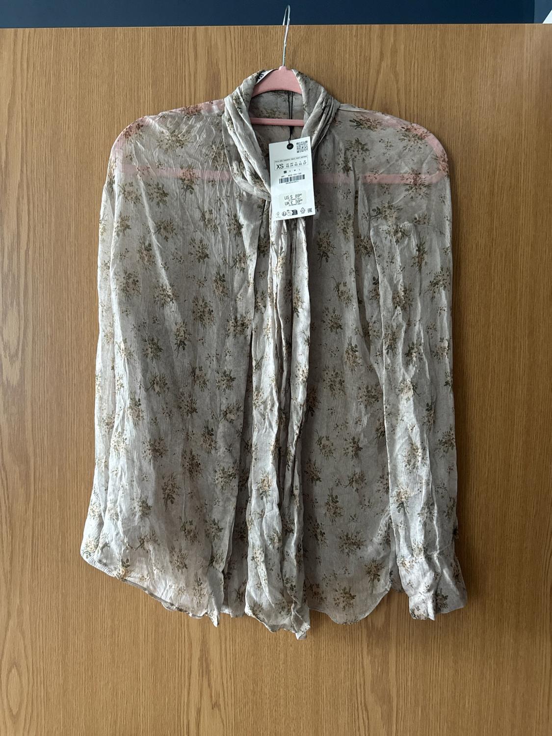 Semi-sheer floral print blouse with a necktie and visible label, hanging on a pink hanger over a wooden door.