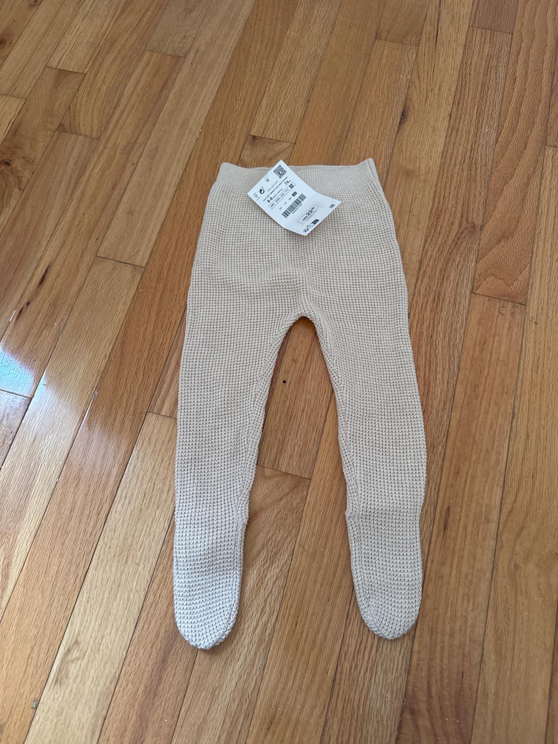 Beige knitted baby trousers with tag, on light wooden floor.