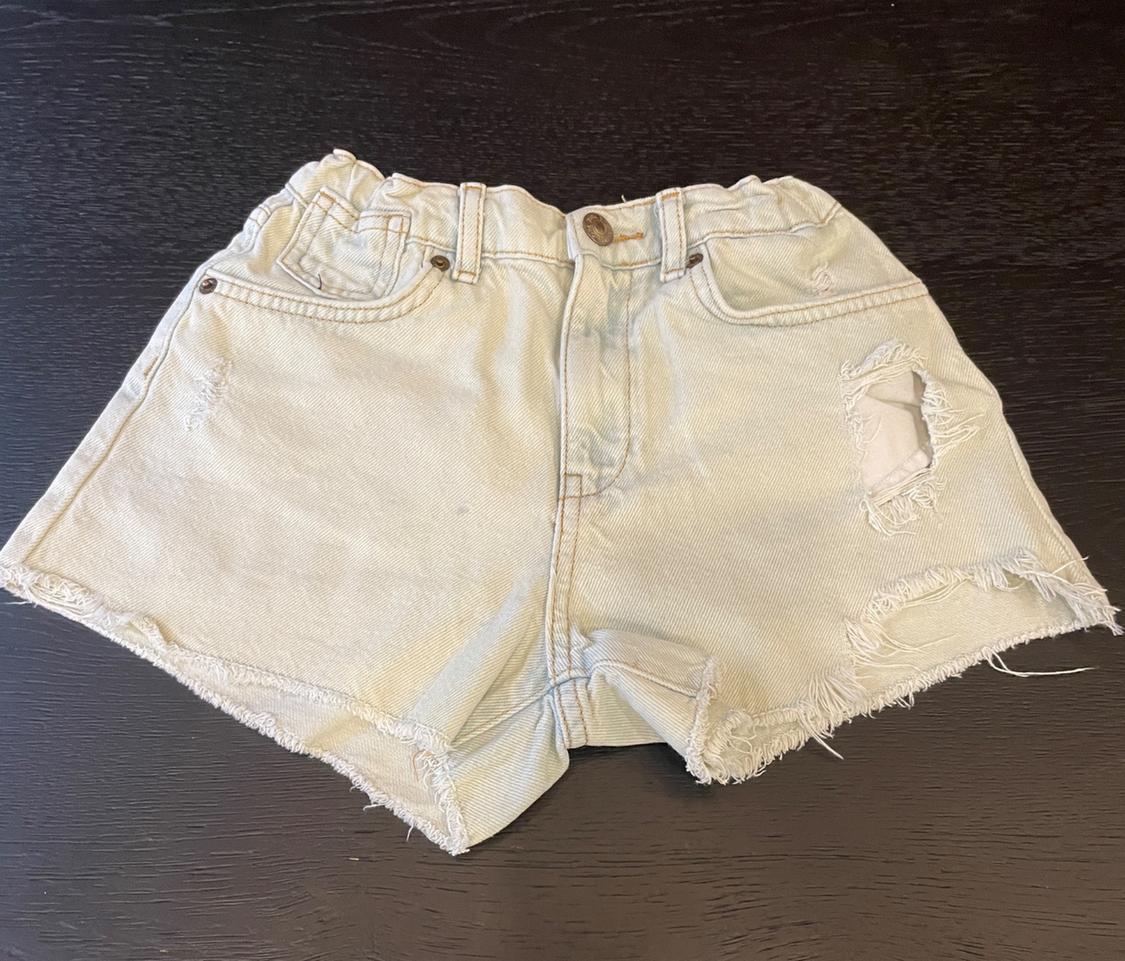 Light wash denim shorts with an elasticated waistband, ripped details and frayed hem, on a dark wood surface.