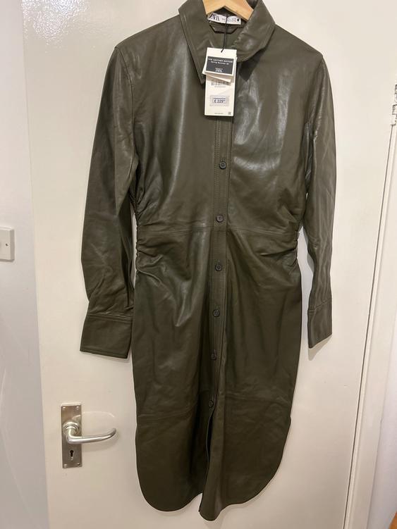 Olive green long coat hanging on a white door, with visible tags on the collar.