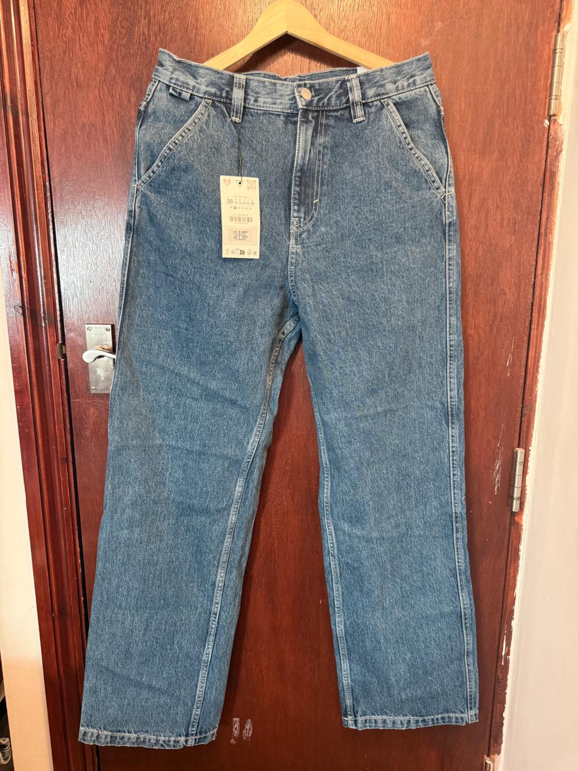 Blue denim jeans hanging on a wooden hanger over a brown door. They have a tag with the number 38. Casual style.