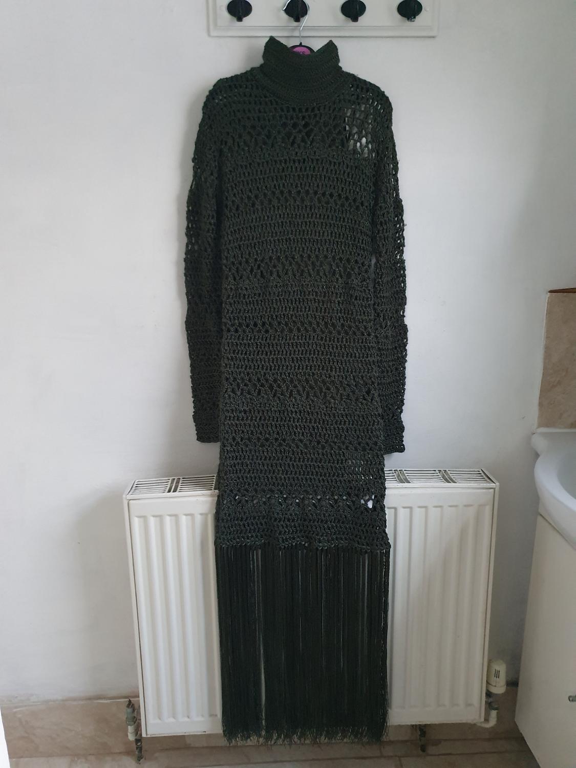 Black knitted dress with a high neck and long fringes hanging on a hanger over a white radiator in a room.