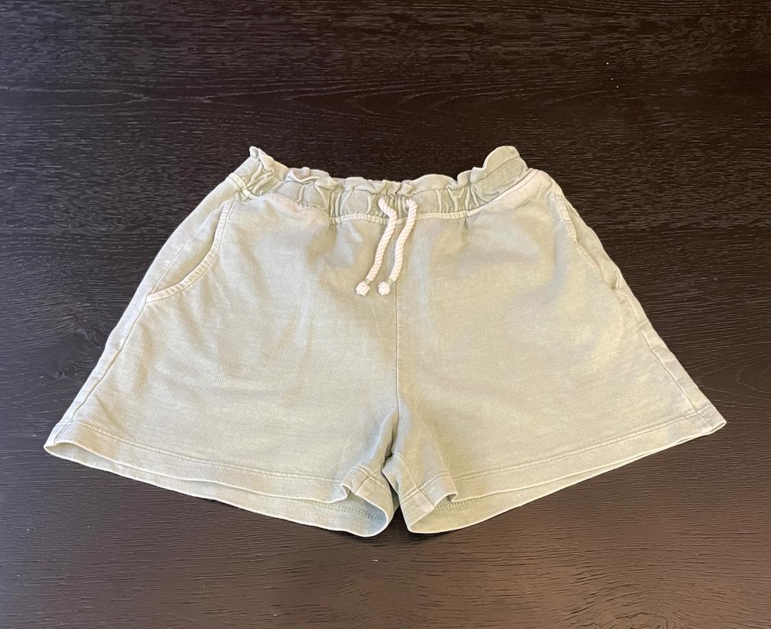 Light green casual shorts with an elasticated gathered waist, white drawstring, and pockets, on a dark wooden surface.