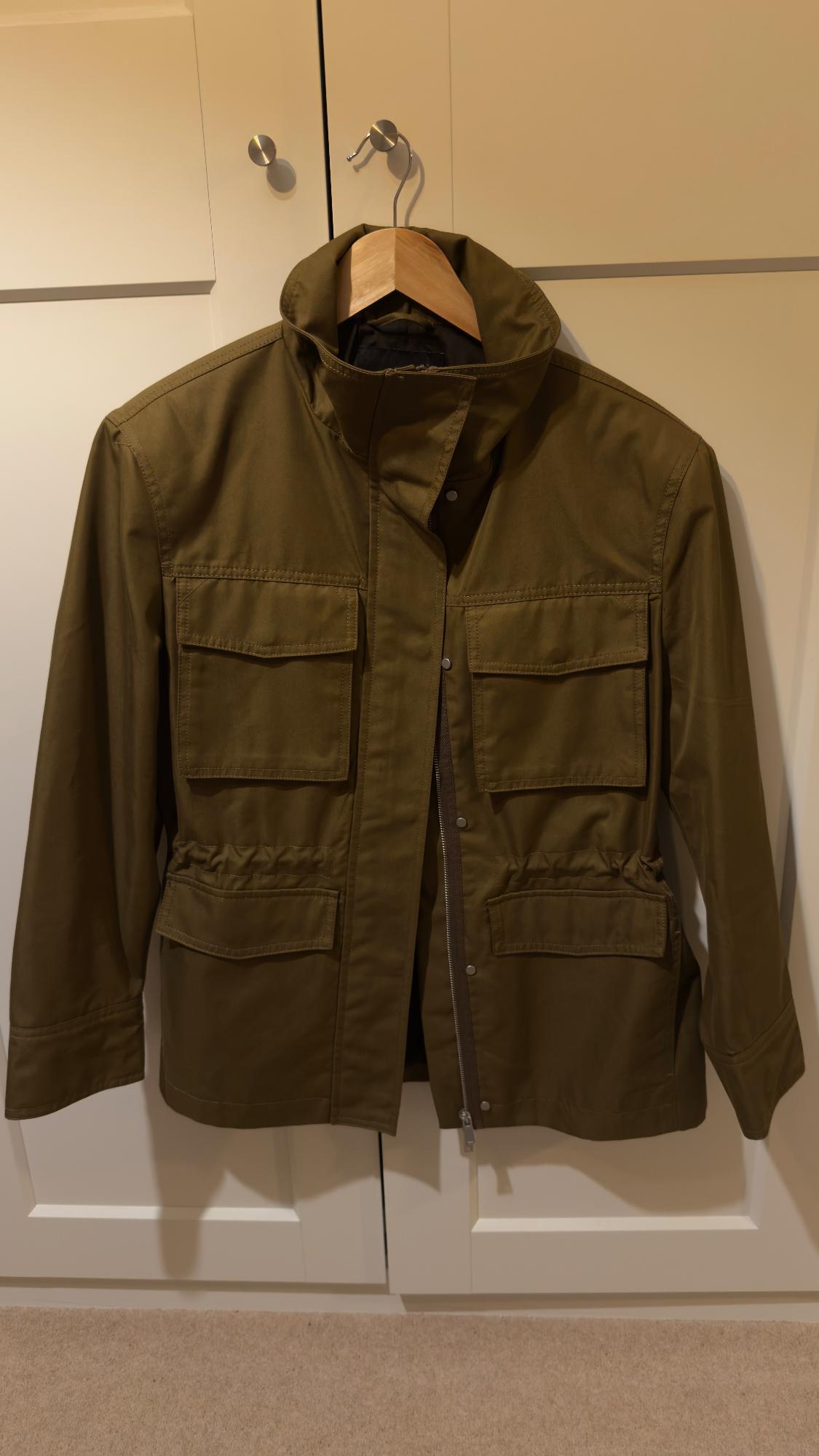 Olive green jacket with large pockets hanging on a wooden hanger in front of a white wardrobe.
