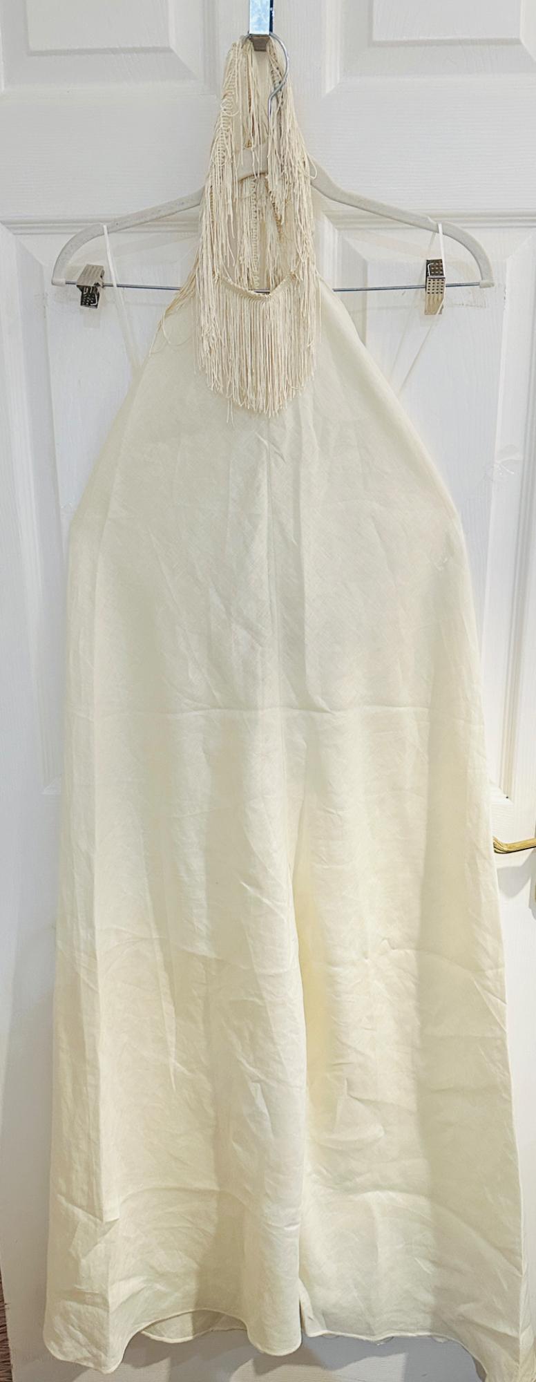 Cream-coloured, relaxed-fit long garment with a halter neck and fringing, hanging on a hanger against a white door.