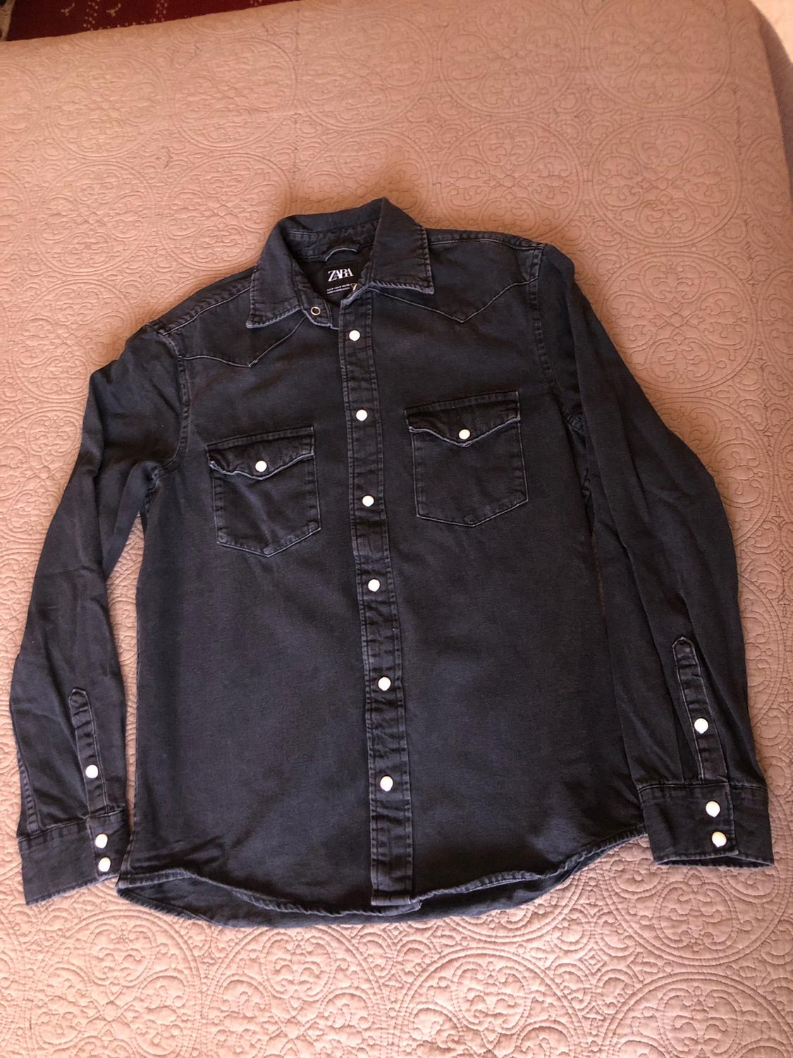 Black long-sleeved shirt with white buttons and two front pockets, on a bedspread with a circular pattern design.