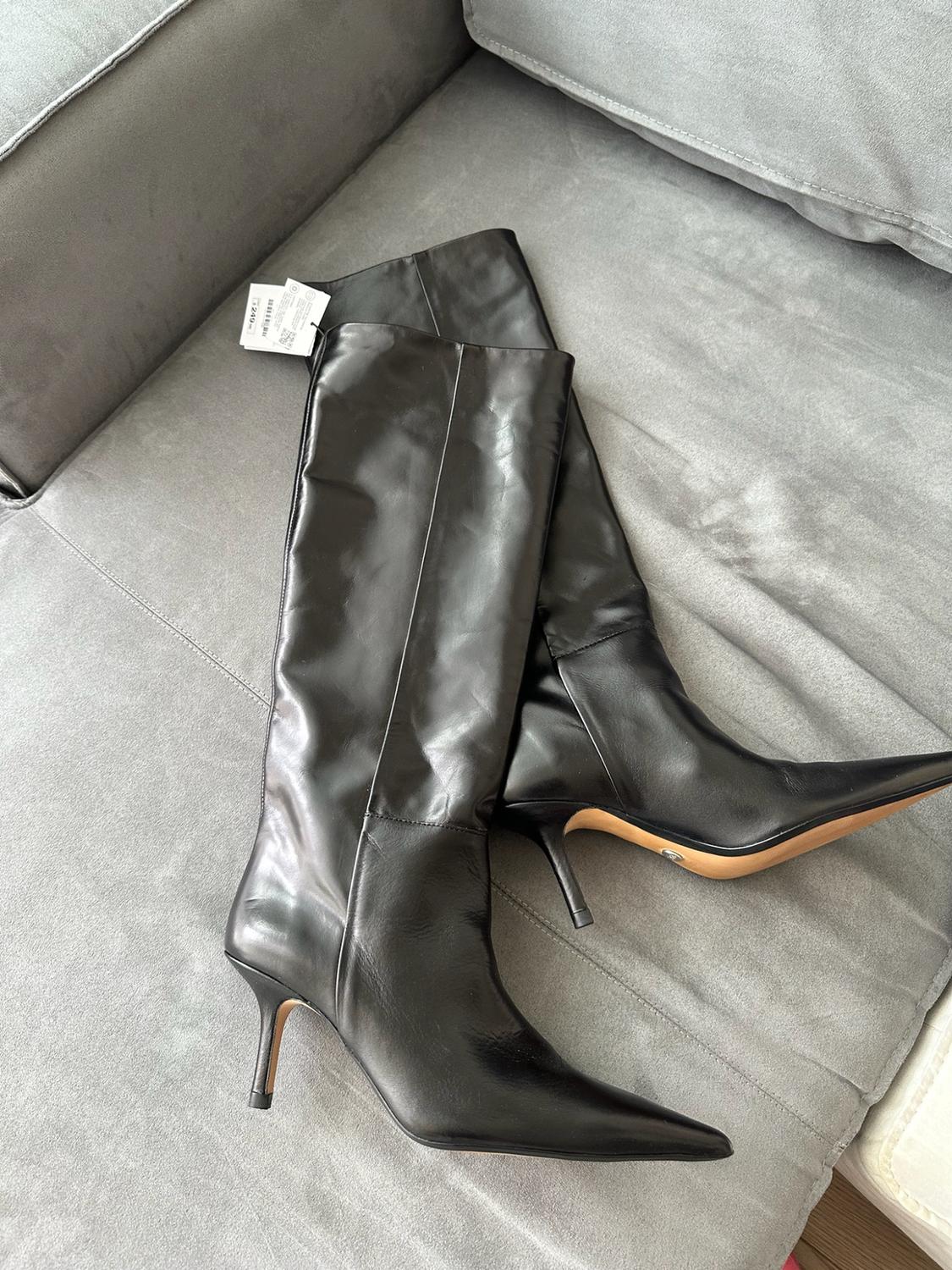 Pair of black high-heeled, pointed-toe boots on a grey sofa. One of them has a white label.