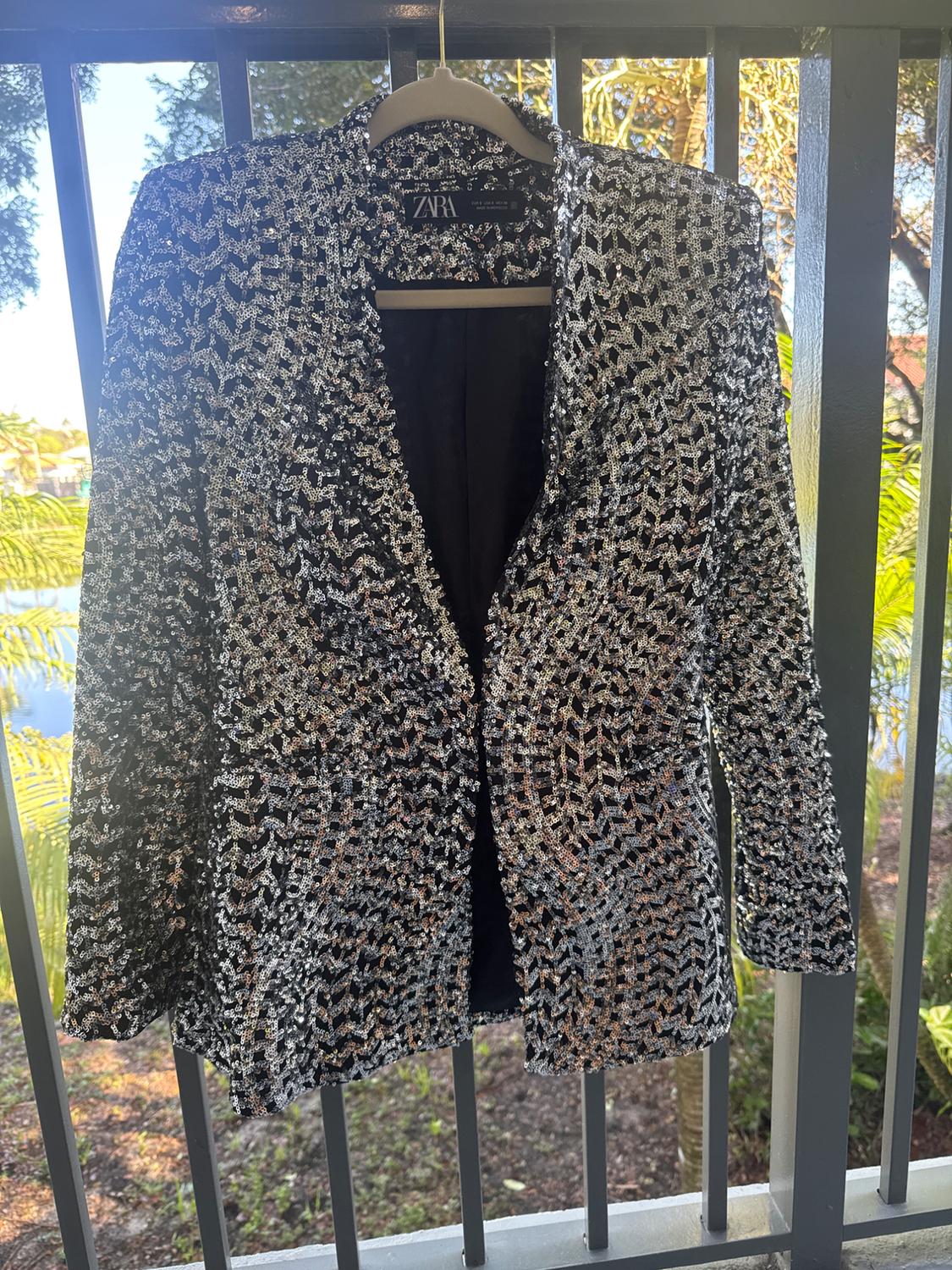Silver and black sequin jacket hanging on a balcony with a vegetation background. Visible tag: ZARA.
