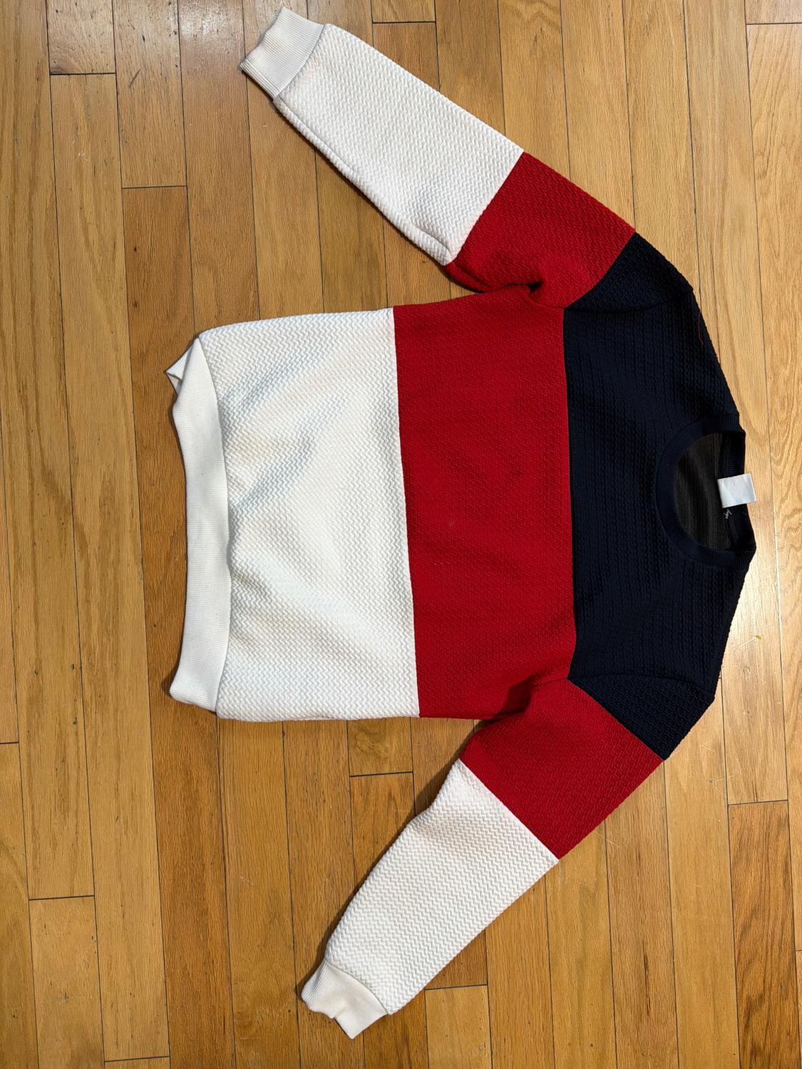 Blue, red, and white color block sweater on a wooden floor. Long-sleeve design with a woven texture.