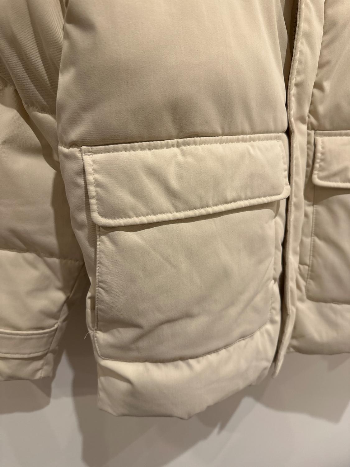 Detail of a beige padded jacket with large pockets and visible stitching, hanging against a white background.