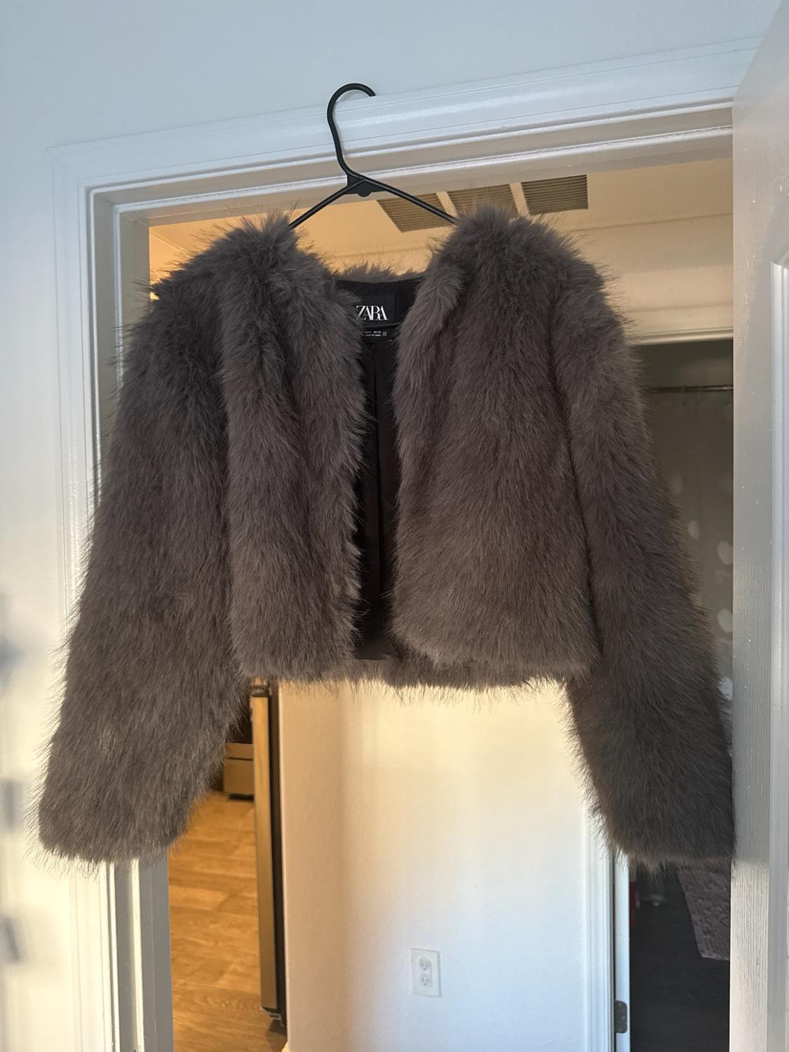 Short grey fur jacket hanging on a hanger on a white door in a domestic interior. Label with the text ZARA.