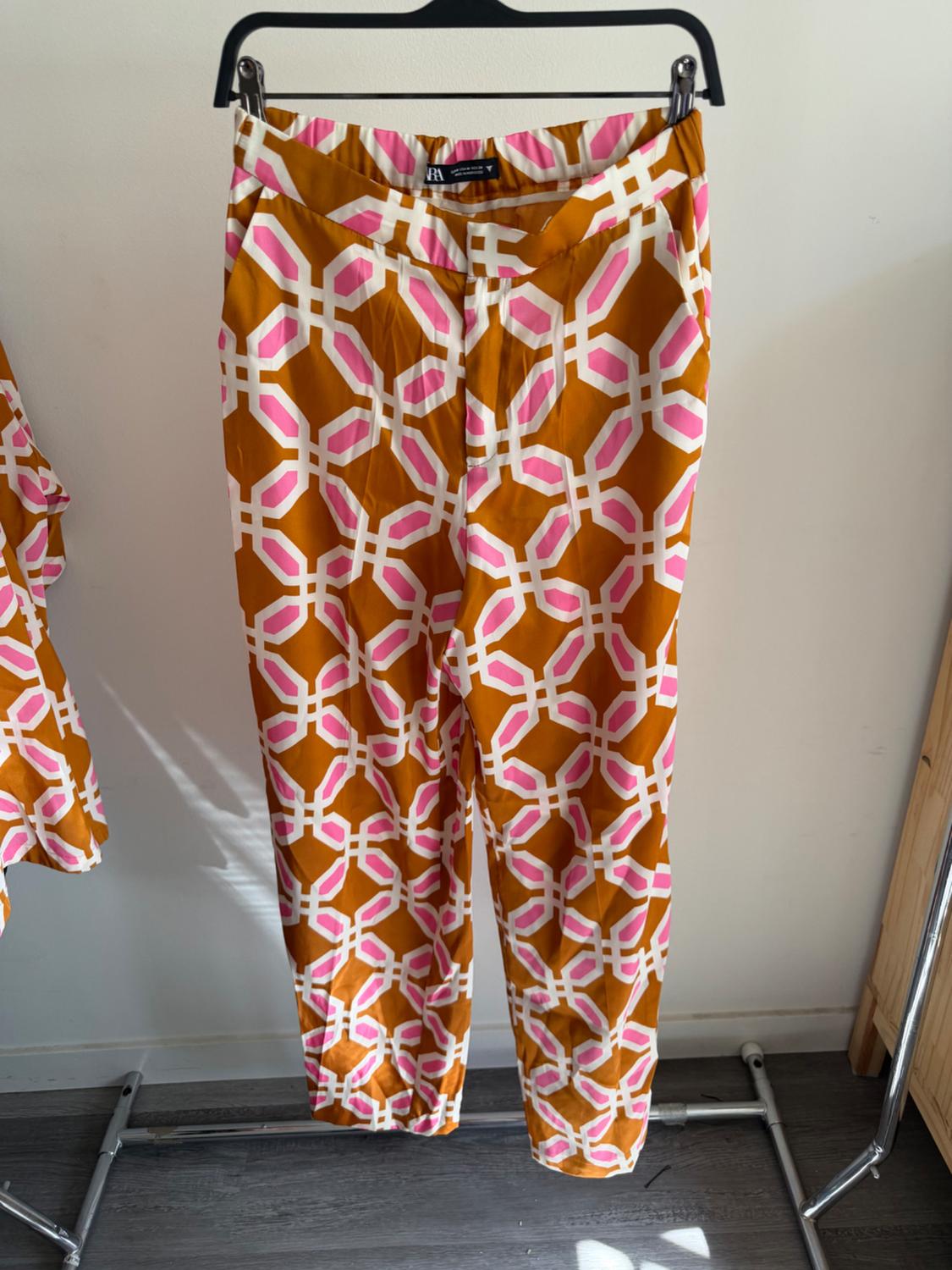 Trousers hung with a geometric print in orange, white, and pink tones. White wall background and grey wooden floor.