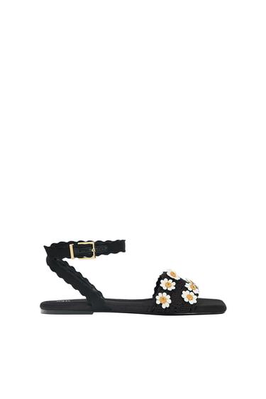 Black flat sandals with ankle strap, gold buckle and front strap embellished with white and yellow flowers on a white background.