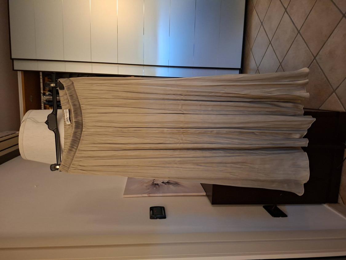 Long pleated beige skirt hanging on a black hanger, with a white wardrobe background and tiled floor.