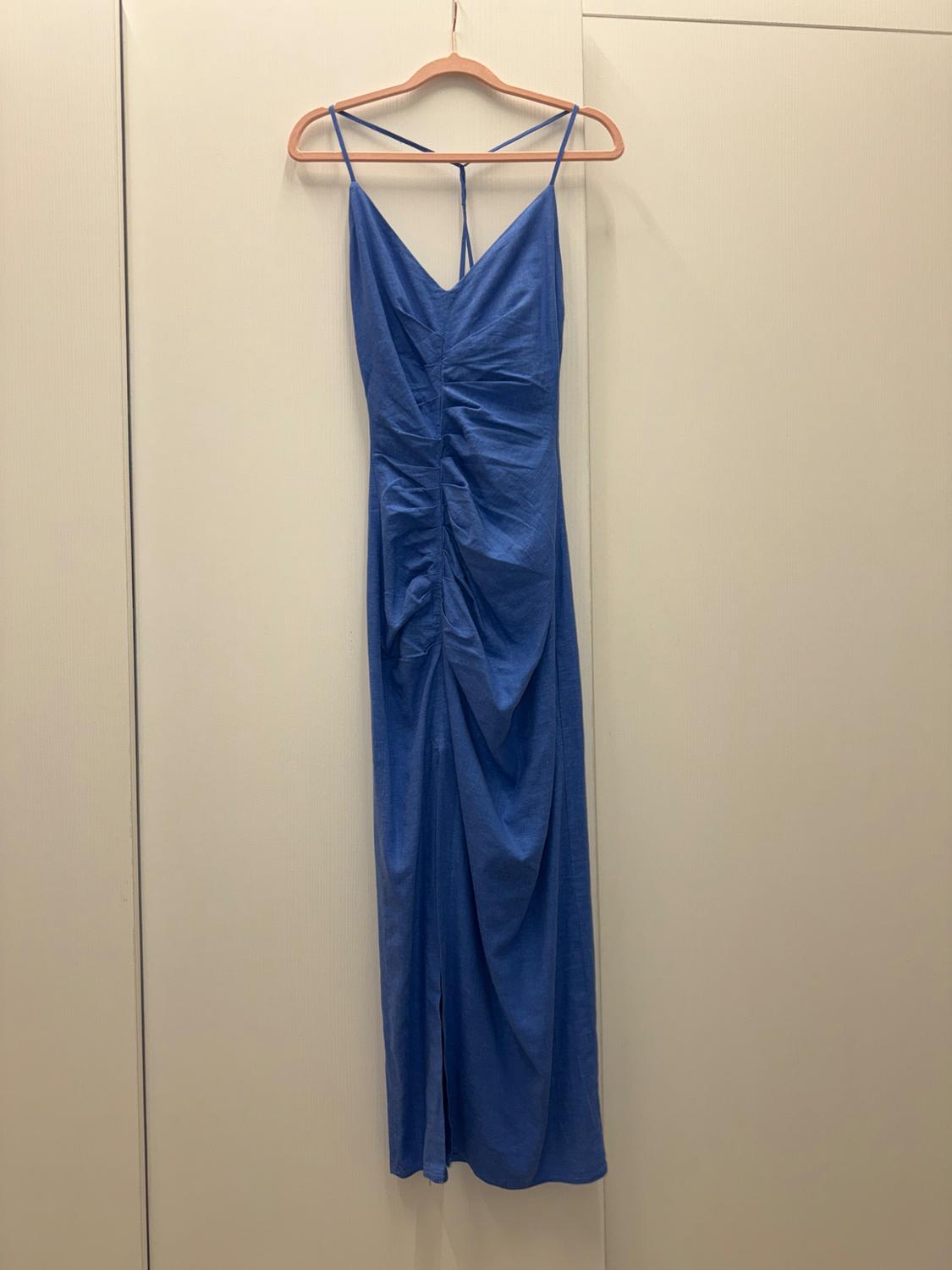 Blue dress with thin straps and central ruching, hanging on a wooden hanger against a beige background.