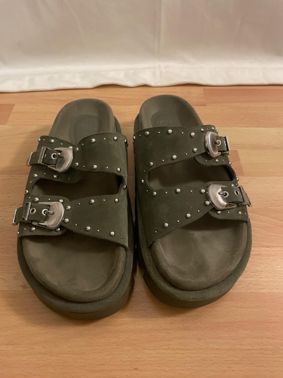 Green sandals with studs and metal buckles on a light wooden floor, white background.