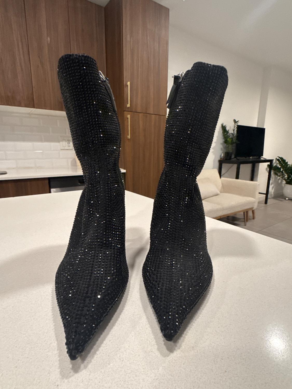 Black high-heeled, pointed-toe boots, covered in glitter, on a white table in a modern room with wooden furniture.