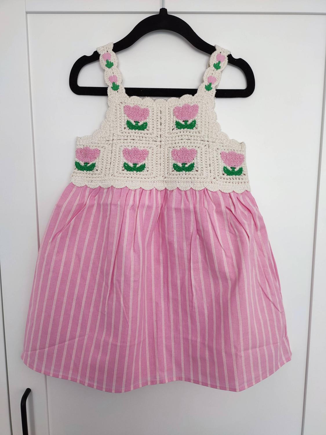 Dress hanging on a black hanger on a white door. Beige crochet bodice with pink flowers and a pink and white striped skirt.