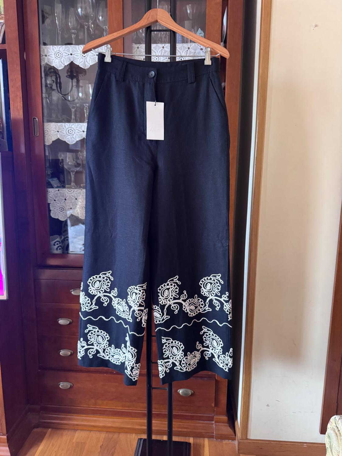 Black trousers hanging on a hanger, with white embroidery on the hem. Wooden furniture background with crystal glasses.