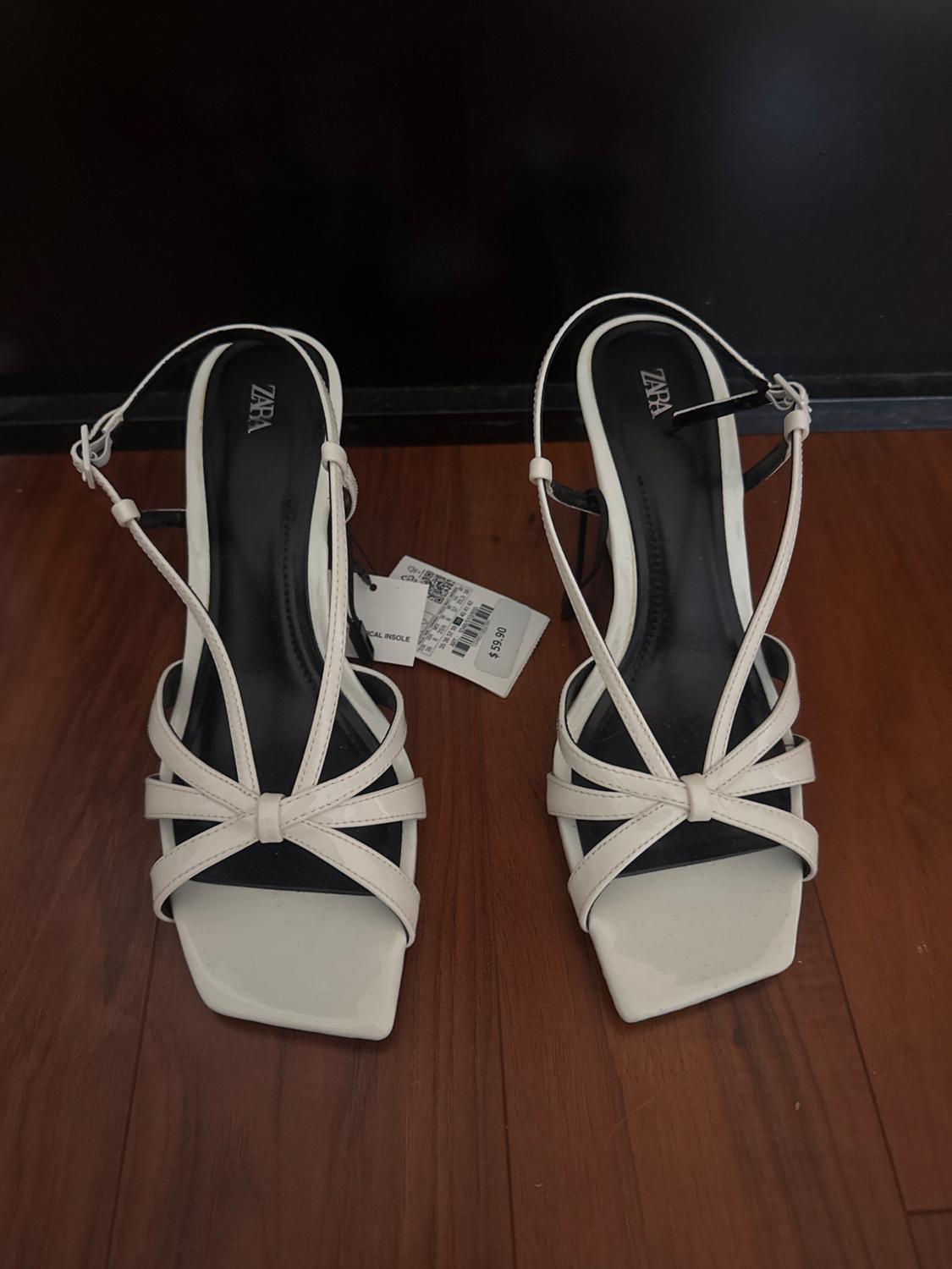 White cross-strap sandals with a block heel on a wooden floor. Visible label with the text "ZARA".