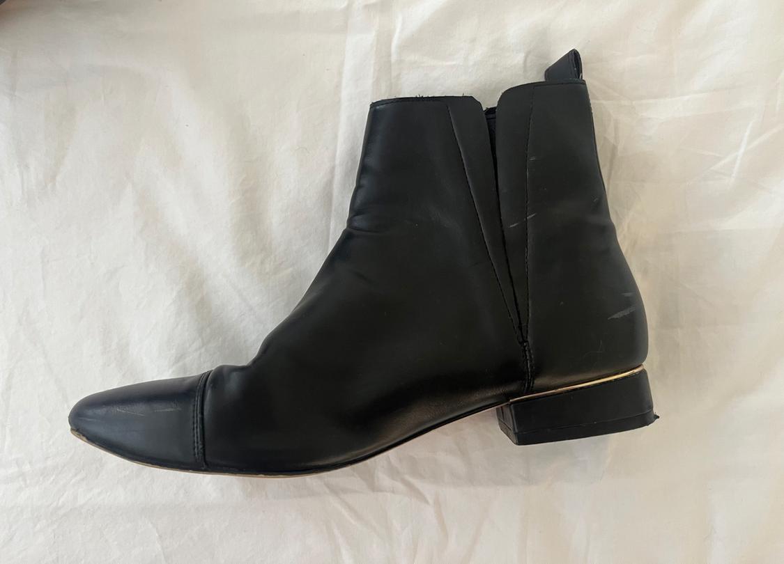 Plain black low-heeled ankle boot with gold detail on the heel, set against a crumpled white fabric background.