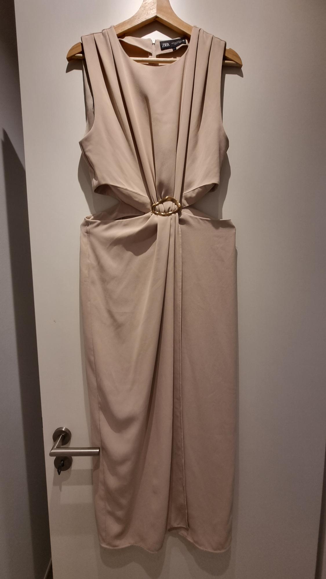 Sleeveless beige dress hanging on a wooden hanger, with a gold ring detail at the waist. White door background.
